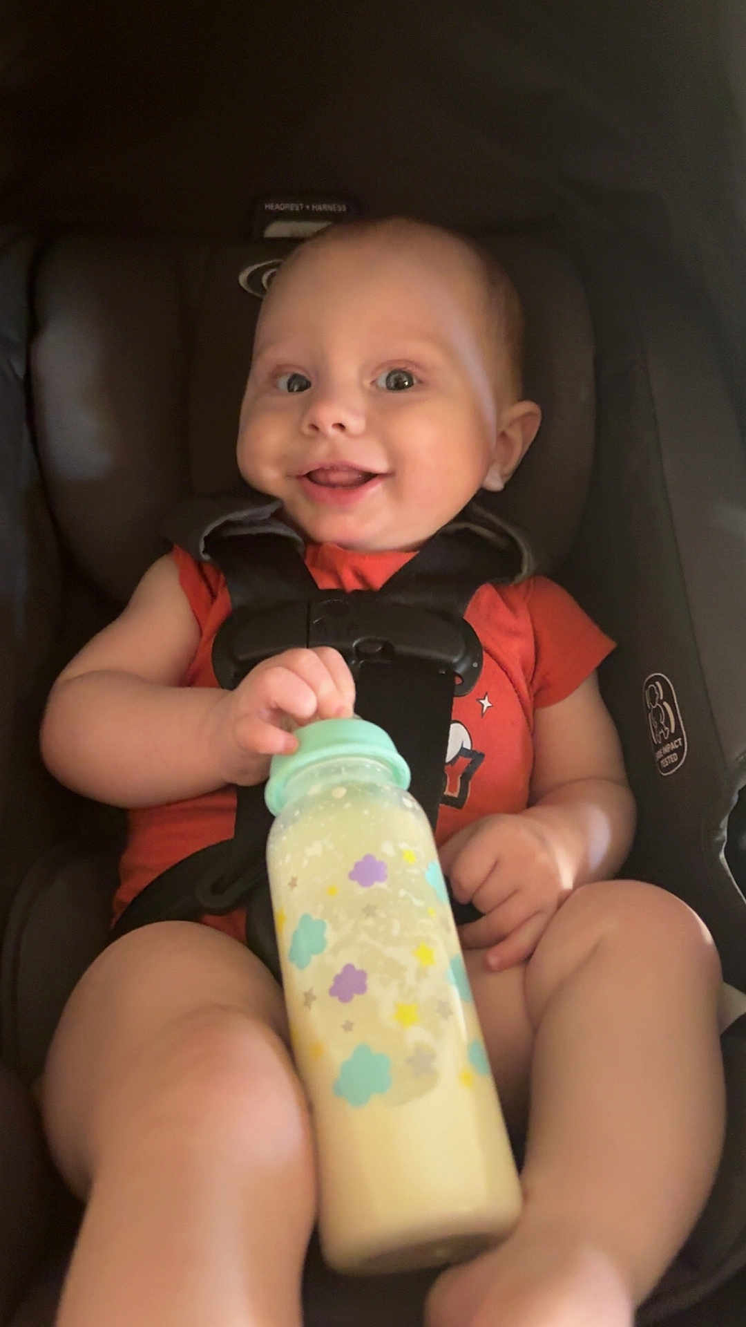 Kalix joined the competition — help win amazing prizes! baby, infant, car_seat, bottle, milk, smiling, happy, child, seatbelt, orange_shirt, cute, portrait, young_child, indoors, holding, feeding, baby_bottle, sitting, face, person
