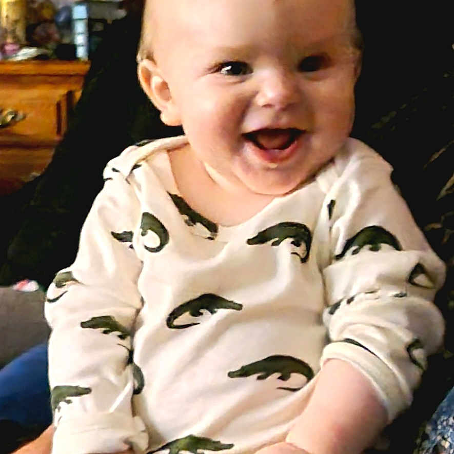 Caio is registered to the contest to win money with this photo: alligator_pattern, baby, child, clothing, cozy, cute, expression, face, furniture, hand, happy, indoor, infant, onesie, person, phone, portrait, seated, skin, smiling