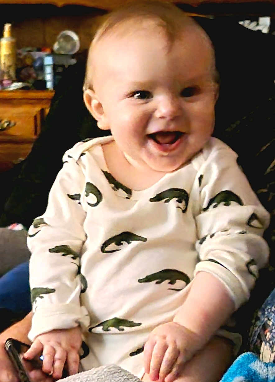 Caio is registered to the contest to win money with this photo: baby, smiling, child, onesie, alligator_pattern, indoor, person, happy, cute, seated, hand, face, infant, clothing, phone, furniture, cozy, portrait, skin, expression