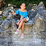 child, girl, outdoor, rocks, stone_bench, blue_top, white_shorts, sneakers, scrapes, sunlight, nature, tree, greenery, smile, casual_clothing, sitting, daylight, young_person, park, relaxing