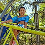 child, girl, playground, climbing, blue_top, white_shorts, yellow_structure, slide, rope_net, outdoor, trees, sunlight, smiling, sneakers, play, recreation, park, daytime, happy, nature