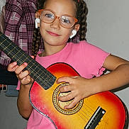 Beatriz participe au concours pour gagner de l'argent avec cette photo : child, girl, glasses, braids, guitar, musical_instrument, earbuds, pink_shirt, indoor, plaid_jacket, wall, posing, smiling, person, music, casual, young, playing, accessory, technology