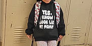Marley is registered to the contest to win money with this photo: child, locker, backpack, hoodie, text_on_clothing, shoes, floor, school, smiling, standing, indoor, person, young_child, patterned_clothing, casual_wear, hair_braids, expression, wall, metal, tile_floor
