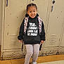 child, locker, backpack, hoodie, text_on_clothing, shoes, floor, school, smiling, standing, indoor, person, young_child, patterned_clothing, casual_wear, hair_braids, expression, wall, metal, tile_floor