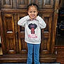 child, girl, smiling, standing, white_sweatshirt, princess_graphic, blue_jeans, silver_shoes, carpet, wooden_cabinet, indoor, portrait, cute, happy, fashion, casual_clothing, person, home, floor, young