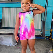 Marley is registered to the contest to win money with this photo: child, girl, standing, peace_sign, colorful_clothing, rainbow, outdoor, concrete, swing, footwear, sandals, smiling, shorts, tank_top, curly_hair, playful, portrait, daylight, background, furniture