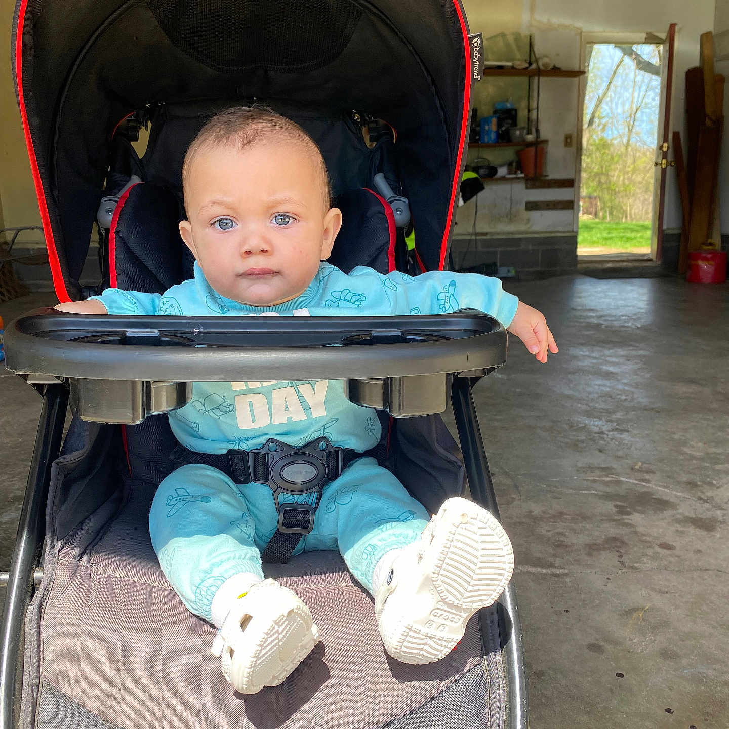 Eziekel joined the competition — help win amazing prizes! baby, baby_boy, blue_eyes, blue_outfit, casual_clothing, child, curious, daylight, expression, floor, garage, indoor, infant, open_door, person, playful, seat, shoes, stroller, sunlight