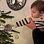 baby, child, christmas_tree, ornament, reflection, hand, striped_sweater, indoor, holiday, decor, mirror, pine_tree, curious, person, face, warm_light, seasonal, celebration, smiling, support