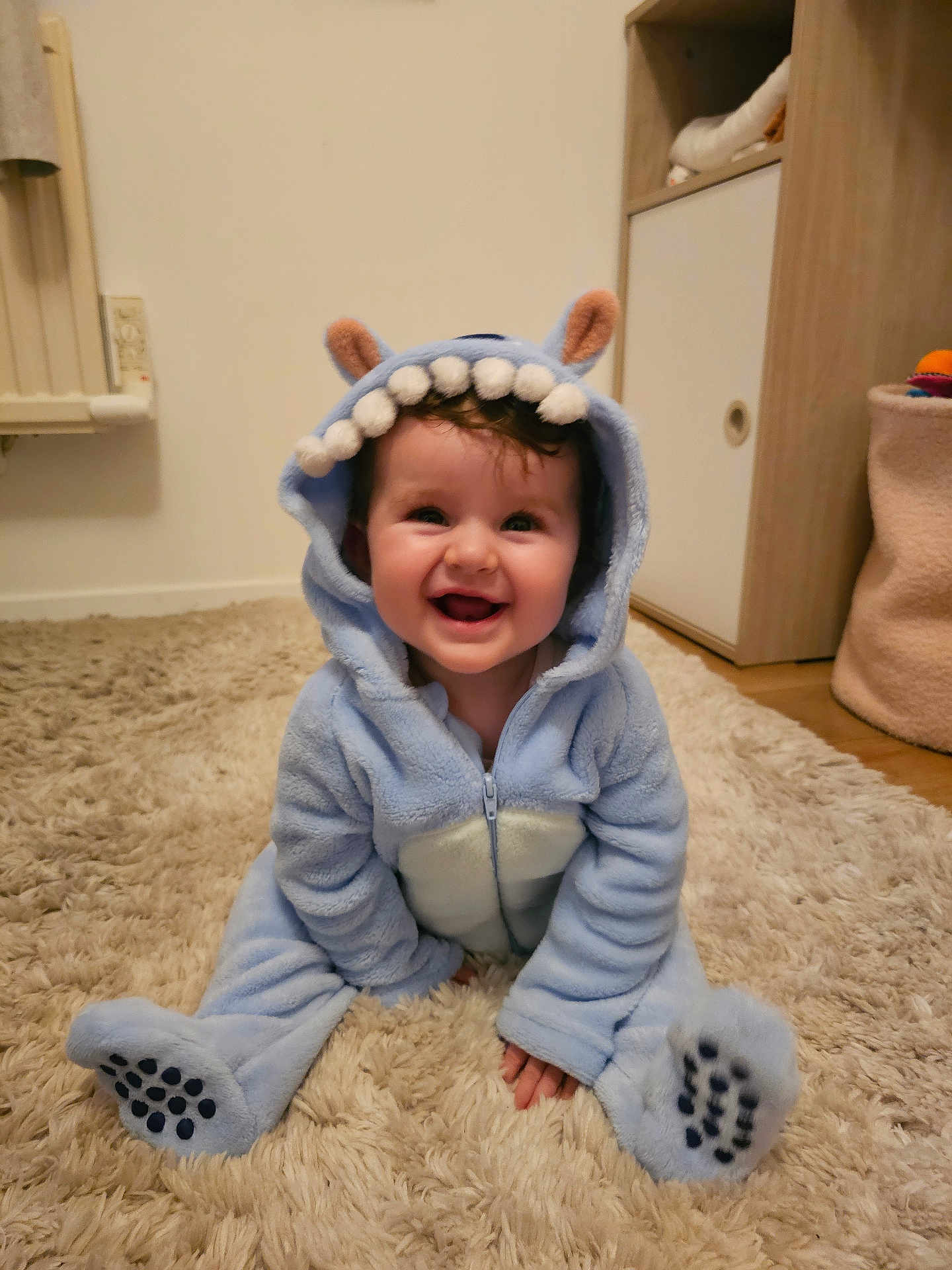 Giulia participe au concours pour gagner de l'argent avec cette photo : baby, child, smiling, blue_onesie, costume, monster_costume, ears, teeth, carpet, floor, indoor, cute, happy, infant, sitting, clothing, person, portrait, room, furniture
