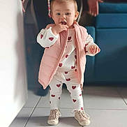 Layana participe au concours pour gagner de l'argent avec cette photo : baby, toddler, child, pink_vest, heart_pattern, white_outfit, shoes, standing, tiled_floor, caregiver_hands, living_room, couch, portrait, cute, facial_expression, hair_ponytail, pacifier_clip, assistance, casual_clothing, indoor