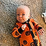 baby, infant, carpet, orange_clothing, onesie, pumpkin_pattern, jack_o_lantern, cute, lying_down, hand, face, expression, indoor, soft_texture, child, person, newborn, black_buttons, smirk, cozy