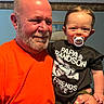Poseidon is registered to the contest to win money with this photo: baby, background, black_clothing, child, expression, face, family, grandfather, hand, holding, home, indoor, man, orange_clothing, pacifier, person, portrait, shirt, smile, toddler
