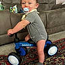 baby, balance_bike, blue, carpet, child, cute, floor, footwear, furniture, gray_clothing, indoor, living_room, pacifier, person, play, rug, smiling, sock, sofa, toy