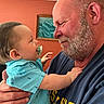 adult, baby, beard, blue_shirt, caring, child, closeup, face, family, gentle, hand, holding, home, indoor, interaction, love, man, pacifier, portrait, smile