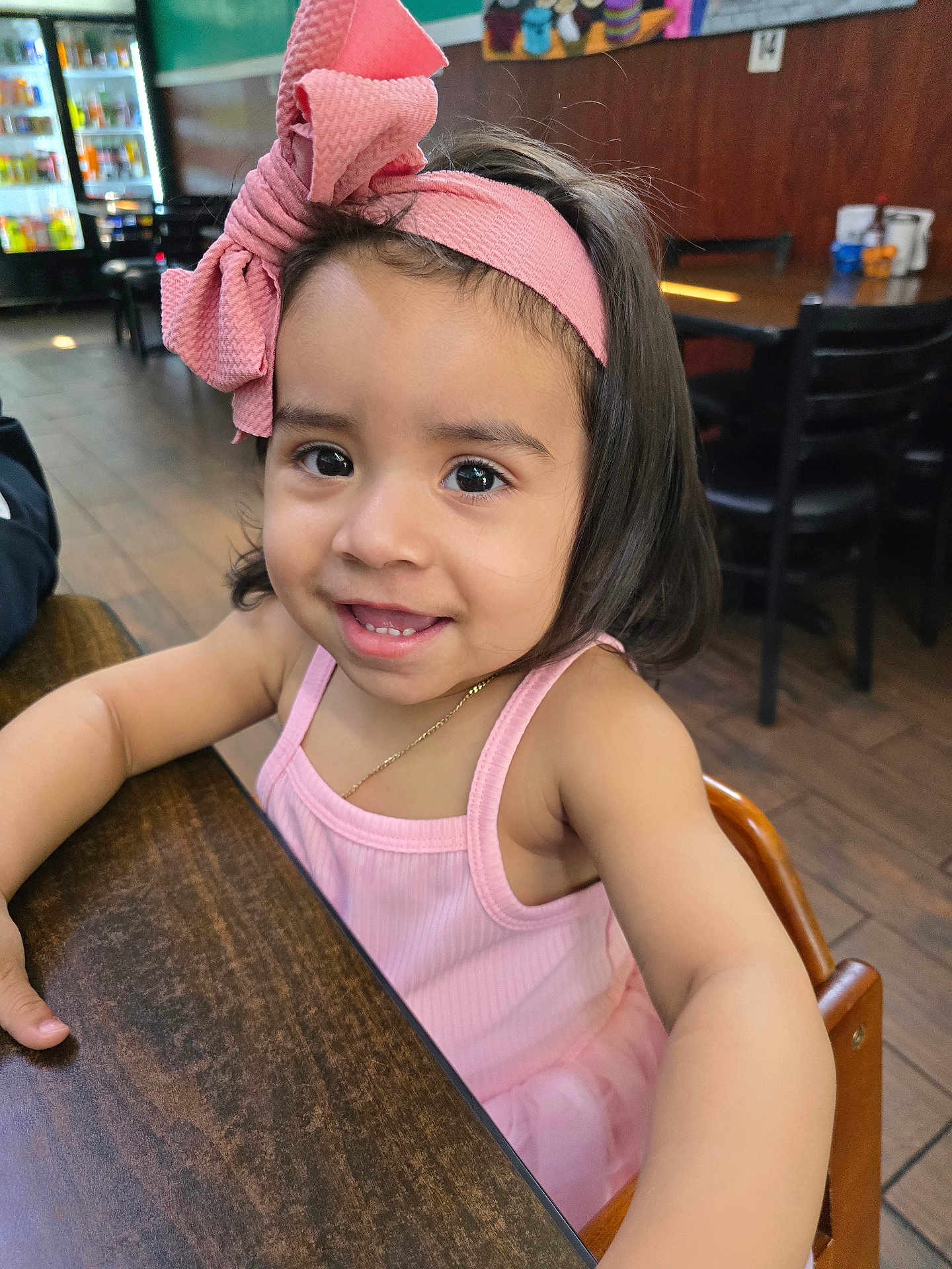 Lily joined the competition — help win amazing prizes! child, toddler, girl, smiling, pink_bow, headband, pink_dress, table, wooden_table, indoor, restaurant, chair, brown_floor, happy, portrait, person, cute, young_child, casual_clothing, seated