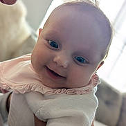 Millie joined the competition — help win amazing prizes! baby, smiling, face, child, infant, person, closeup, portrait, clothing, white, pink, ruffles, hand, adult, holding, indoors, soft_light, blurred_background, cute, happy