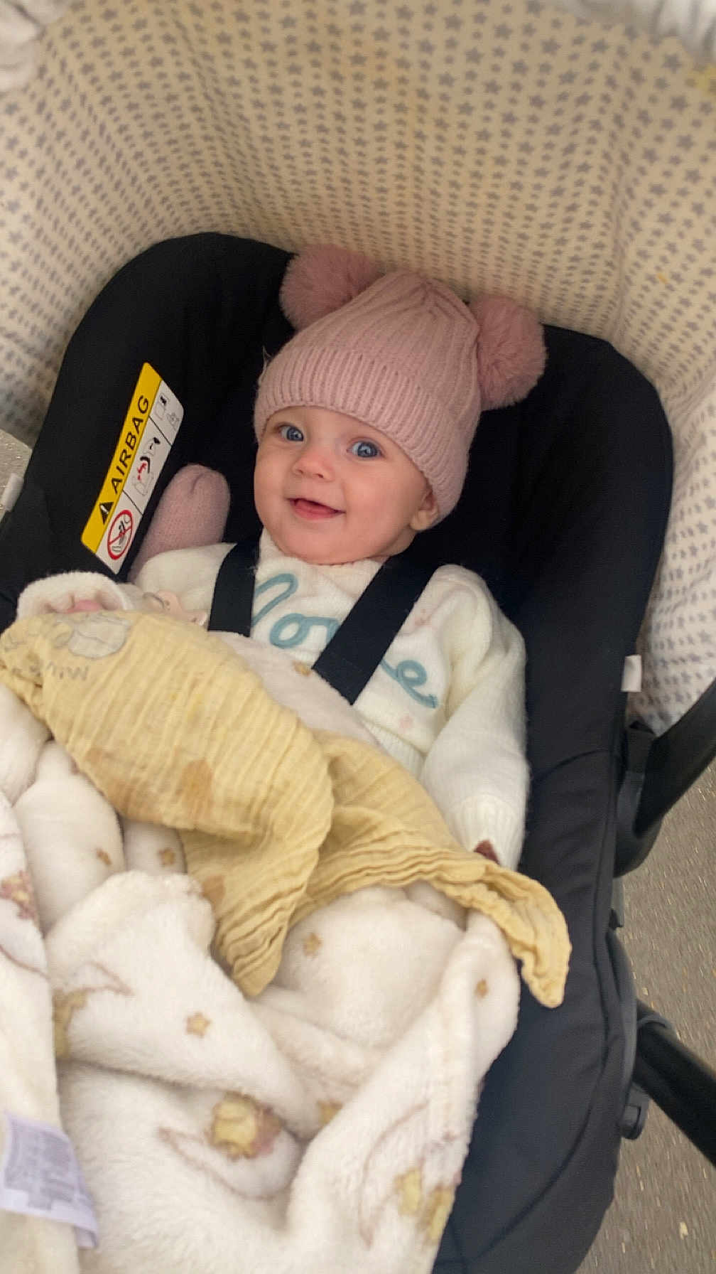 Athena is registered to the contest to win money with this photo: baby, child, smile, hat, pink_hat, blanket, stroller, car_seat, baby_clothing, cozy, cute, pom_poms, happy, infant, face, blue_eyes, indoors, warm, comfort, patterned