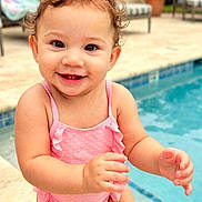 Paisley is registered to the contest to win money with this photo: baby, toddler, child, smiling, happy, poolside, swimming_pool, outdoor, sunny, portrait, curly_hair, pink_swimsuit, standing, arms, hands, tropical_plants, patio, lounge_chair, water, family_photo