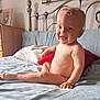 toddler, child, smiling, bed, blanket, pillow, blue_blanket, red_pillow, metal_headboard, wooden_dresser, framed_photo, indoor, happy, skin, person, furniture, room, portrait, sitting, cute
