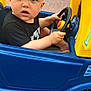 Blake is registered to the contest to win money with this photo: toddler, child, toy_car, steering_wheel, blue_eyes, curious, outdoor, play, plastic, yellow, blue, black_shirt, expression, face, hands, vehicle, wheel, pavement, brick_wall, window