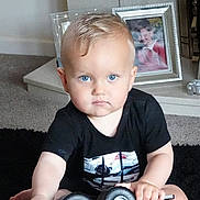 Blake joined the competition — help win amazing prizes! toddler, child, blue_eyes, black_shirt, toy_vehicle, indoor, carpet, living_room, photograph, frame, furniture, person, face, sitting, young_child, short_hair, serious_expression, hand, floor, home