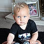 toddler, child, blue_eyes, black_shirt, toy_vehicle, indoor, carpet, living_room, photograph, frame, furniture, person, face, sitting, young_child, short_hair, serious_expression, hand, floor, home