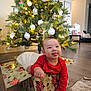 Thomas is registered to the contest to win money with this photo: baby, child, christmas_tree, gift_box, holiday, red_clothing, indoors, smiling, tongue_out, decorations, ornaments, pine_tree, floor, carpet, living_room, present, festive, cute, happy, home