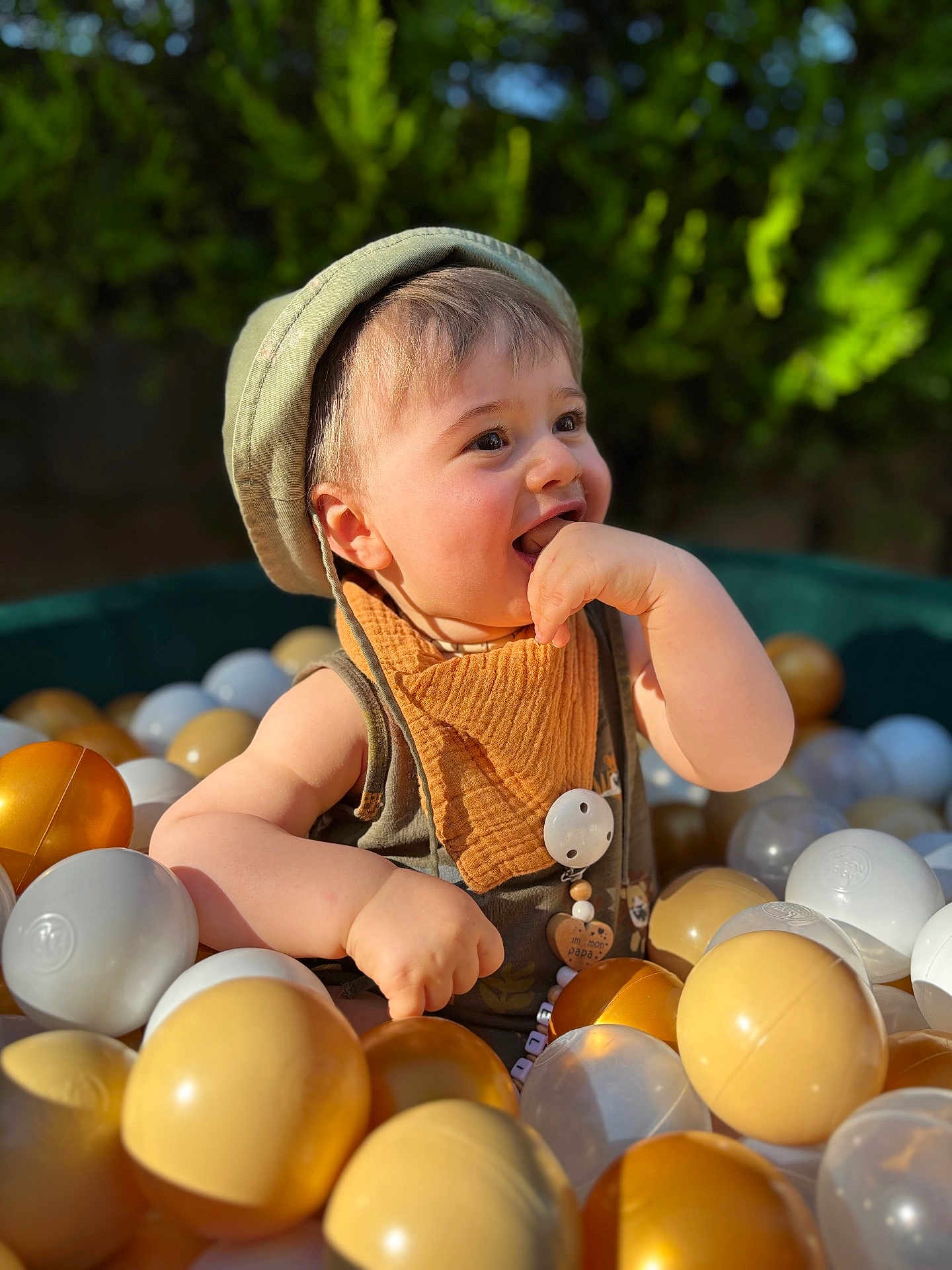 Elio a rejoint le concours — aidez-le/la à gagner de superbes lots ! toddler, child, ball_pit, hat, bib, plastic_balls, outdoor, happy, smiling, sunlight, greenery, play, cute, baby, portrait, cheerful, summer, fun, closeup, colorful