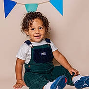 Easton joined the competition — help win amazing prizes! child, toddler, smiling, curly_hair, overalls, white_shirt, sneakers, blue_shoes, indoor, studio, decorations, pennants, happy, sitting, cute, portrait, fashion, feet, clothing, playful