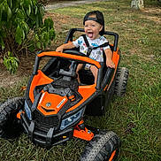 Colton is registered to the contest to win money with this photo: toddler, child, toy_car, outdoor, grass, trees, cap, smiling, playful, happy, nature, vehicle, fun, summer, greenery, young_child, recreation, daytime, expression, casual_clothing