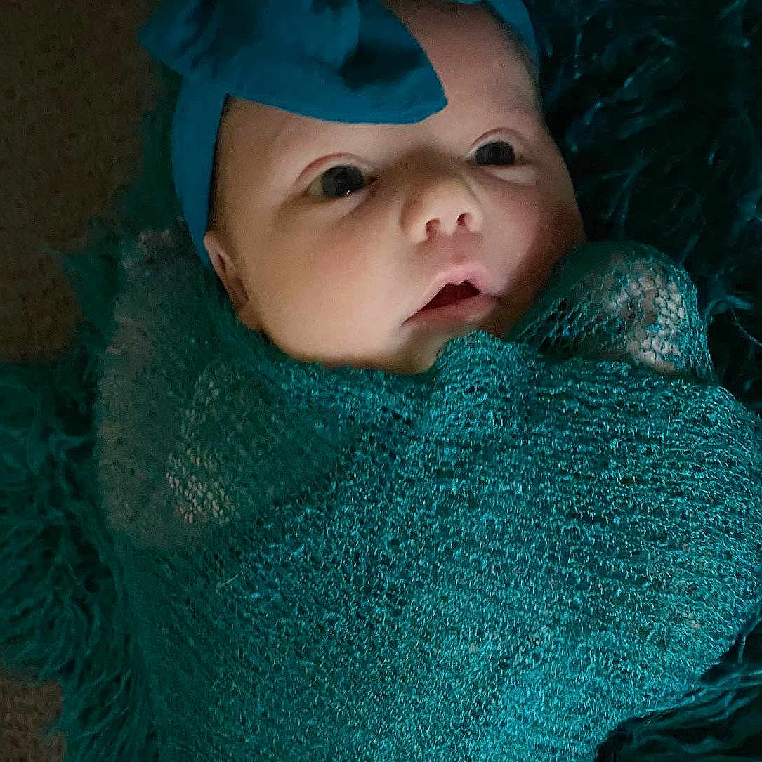 Olivia is registered to the contest to win money with this photo: aqua, azure, baby, baby_toddler_clothing, blue, cap, electric_blue, eye, eyelash, fashion_accessory, headgear, knit_cap, linens, outerwear, pattern, shawl, textile, toddler, wool, woolen
