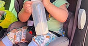 Mateo is registered to the contest to win money with this photo: baby, car_seat, bottle, milk, child, infant, seatbelt, car_interior, sunlight, clothing, patterned_pants, smiling, happy, sitting, young_child, cute, portrait, daylight, safety, vehicle