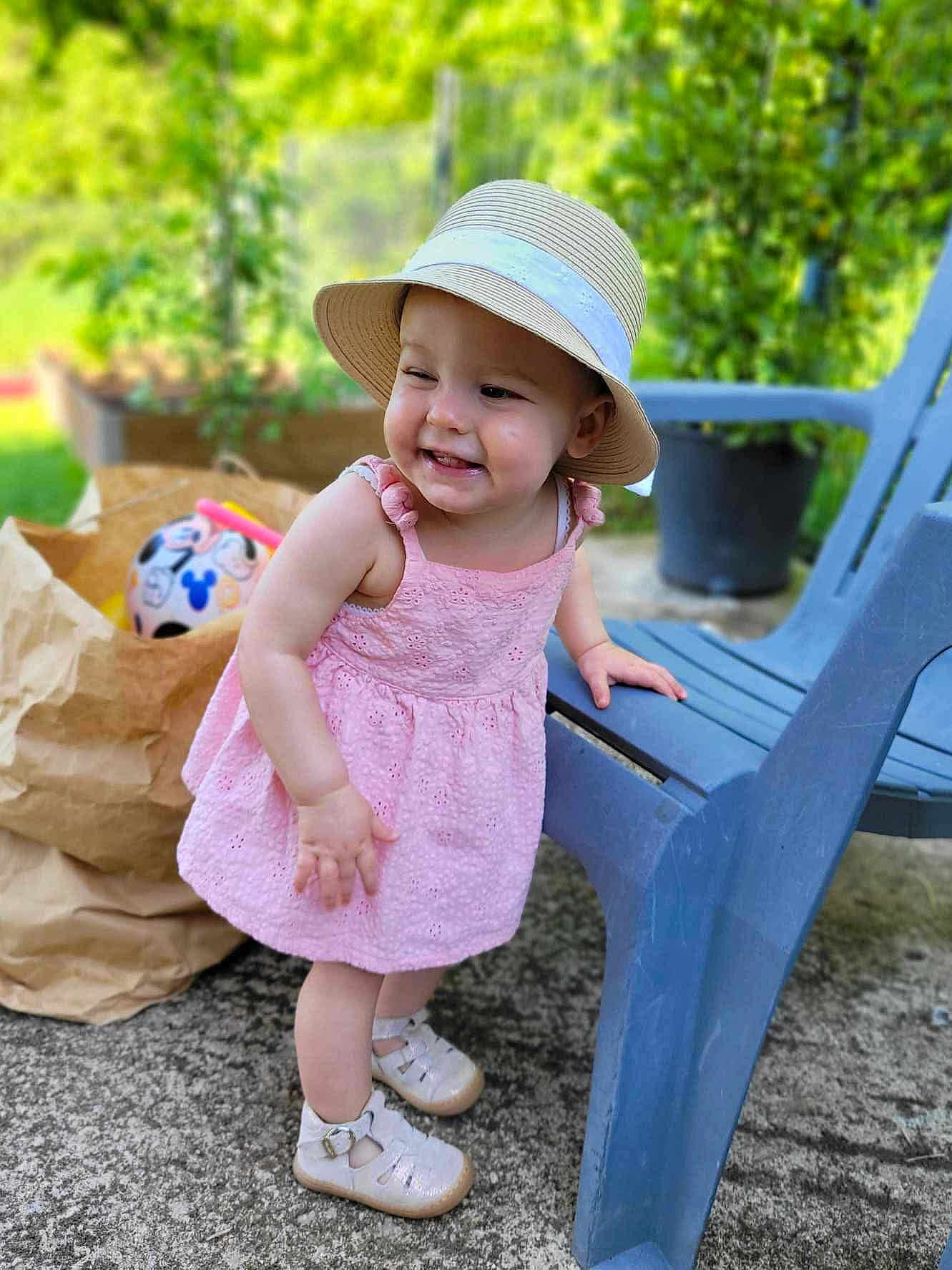 Amalya a rejoint le concours — aidez-le/la à gagner de superbes lots ! toddler, child, pink_dress, hat, smiling, outdoor, blue_chair, paper_bag, ball, greenery, concrete_floor, footwear, happy, playful, sun_hat, summer, baby, cute, daylight, portrait