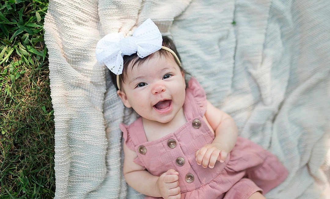 Aniyah is registered to the contest to win money with this photo: baby, child, smiling, happy, outdoor, grass, blanket, headband, bow, pink_dress, cute, infant, portrait, face, skin, eyes, hands, knitted_blanket, lying_down, summer