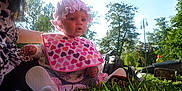 Bonnie is registered to the contest to win money with this photo: baby, child, outdoor, grass, bib, bonnet, person, camera, bottle, trees, sunlight, park, clothing, nature, greenery, sitting, headwear, accessory, daylight, young