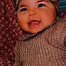 Ahytana participe au concours pour gagner de l'argent avec cette photo : baby, child, smiling, headband, bow, knitted_sweater, cozy, happy, face, portrait, indoor, red_background, patterned_background, cute, infant, person, young_child, warm_clothing, sitting, cheerful