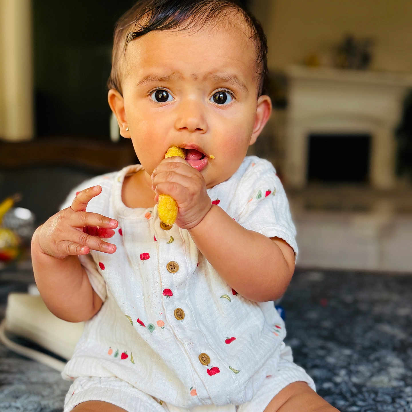Aagya is registered to the contest to win money with this photo: baby, child, snack, cheesy_puff, onesie, indoor, curious, sitting, patterned_surface, face, expression, hand, food, portrait, infant, cute, clothing, young_child, person, furniture
