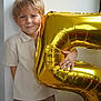 child, boy, balloon, gold, number_five, celebration, smile, blonde_hair, casual_clothing, white_wall, indoor, portrait, happy, hand, light, shadow, young, cute, fun, party