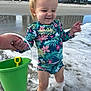 child, toddler, beach, ocean, waves, sand, swimsuit, floral_pattern, bucket, shovel, parent_hand, holding_hands, earrings, splashing, wet_sand, reflection, building, vacation, smile, portrait