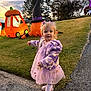 child, girl, purple_jacket, pink_dress, white_shoes, sidewalk, grass, inflatable_decorations, pumpkin, witch_hat, halloween, outdoor, evening, trees, smiling, pointing, cute, holiday, costume, young