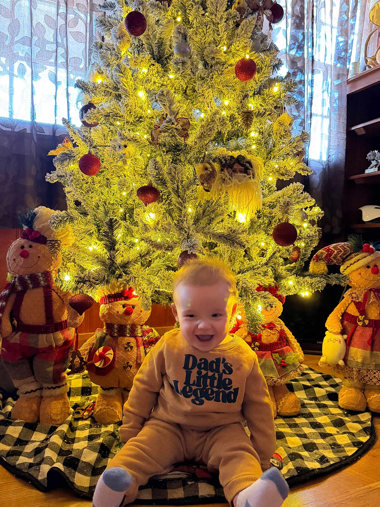 Hudson is registered to the contest to win money with this photo: baby, child, infant, christmas_tree, christmas_lights, ornaments, plush_toy, snowman, holiday, smile, sitting, onesie, tree_skirt, cozy, warm_lighting, festive, curtains, home_interior, decorations, floor