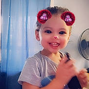London is registered to the contest to win money with this photo: toddler, child, smiling, portrait, indoors, digital_filter, sparkles, red_hair_buns, phone, handheld_device, blue_curtain, fan, seated, short_sleeves, t_shirt, happy, face, eyes, home, photography