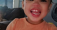 London is registered to the contest to win money with this photo: child, close_up, couch, curtains, eyelashes, face_filter, fan, girl, happy, hearts, home, indoors, laughing, orange_shirt, portrait, seated, smiling, teeth, toddler, unicorn