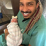Iris joined the competition — help win amazing prizes! man, newborn, baby, hospital, smiling, dreadlocks, tattoos, piercings, blanket, headband, chair, medical_scrubs, person, indoor, happy, seated, portrait, skin, arm, closeup