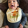 baby, bib, child, clothing, couch, curly_hair, cute, face, giraffe, hand, happy, indoor, infant, pants, person, pillow, portrait, seated, smiling, teddy_bear_pattern