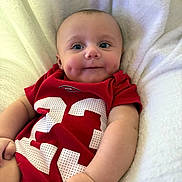 Justice is registered to the contest to win money with this photo: baby, infant, child, face, dimples, blue_eyes, red_onesie, number_23, sports_jersey, blanket, white_blanket, portrait, close_up, adorable, chubby_cheeks, smiling, hands, bedding, cute, human