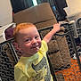 toddler, child, smiling, yellow_shirt, crocodile_graphic, indoor, living_room, safety_gate, wood_floor, recliner_chair, window, playful, red_hair, shorts, person, cute, happy, chewing, handrail, home