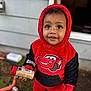 black_clothing, casual_wear, child, close_up, curious, daylight, face, grass, holding_object, hoodie, house_wall, outdoor, person, playful, red_clothing, smiling, toddler, toy_shoes, window, young_child