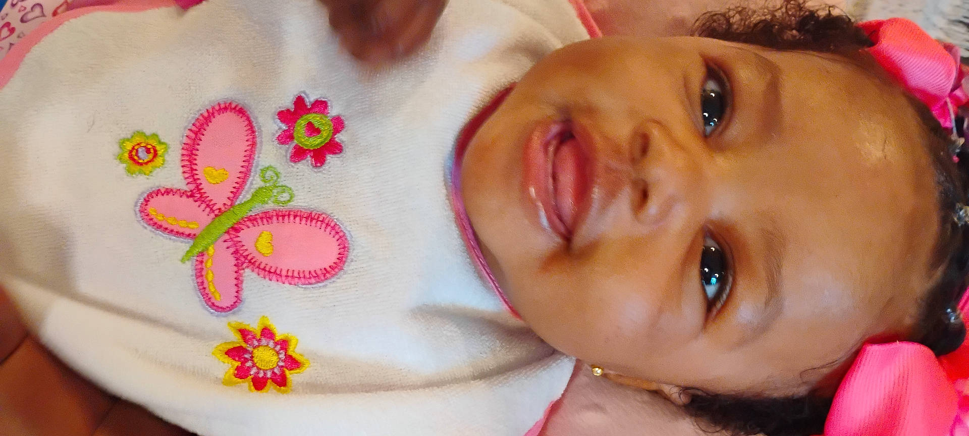 Jehlani is registered to the contest to win money with this photo: baby, bib, butterfly, child, close_up, clothing, cute, embroidery, face, flower, hair_accessory, happy, indoors, infant, person, pink_bow, portrait, skin, smiling, young