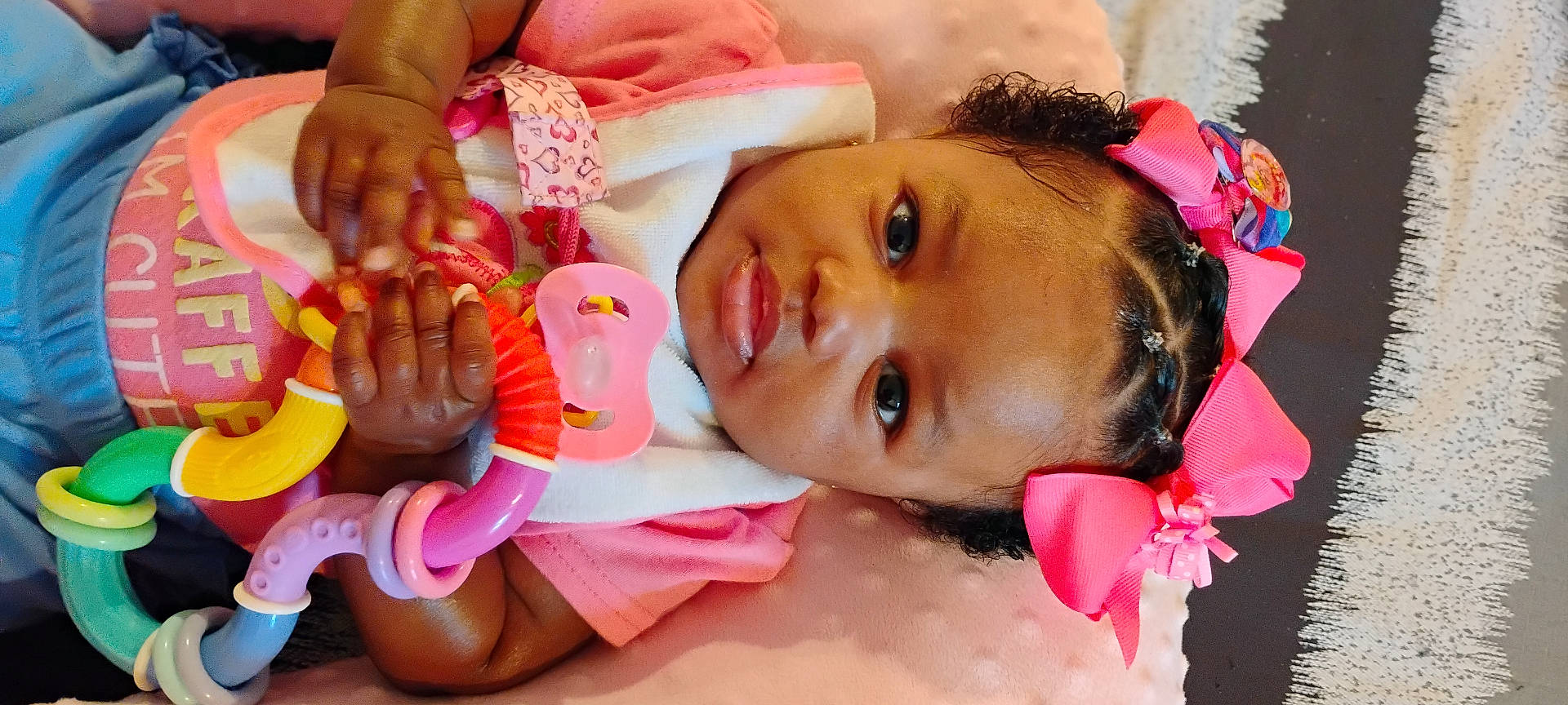 Jehlani joined the competition — help win amazing prizes! baby, bib, blanket, braided_hair, child, closeup, colorful_toy, cute, expression, face, hand, indoor, infant, lying_down, pink_bow, playful, portrait, soft_texture, teething_toy, young_child